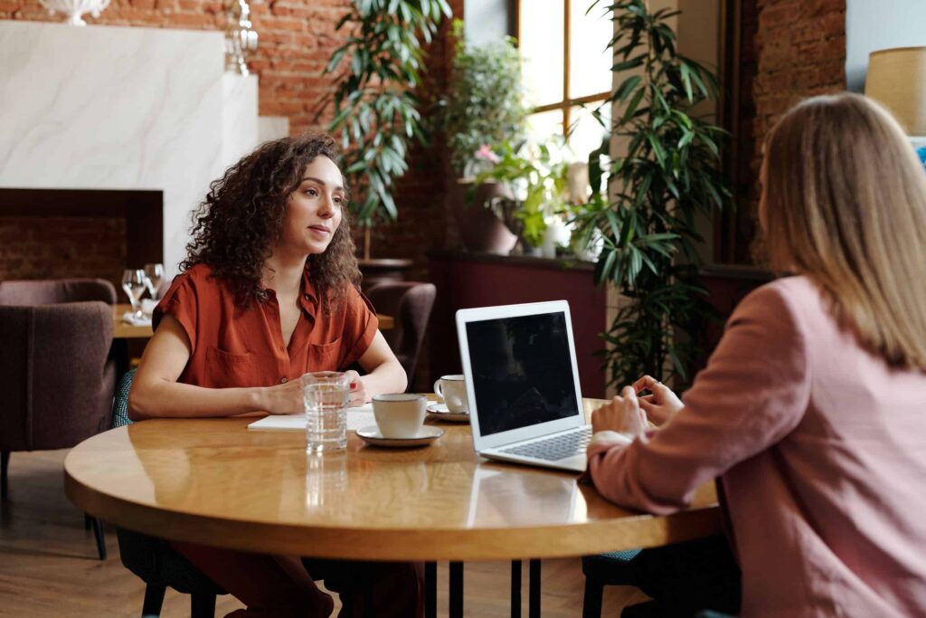 A woman sits at a round table across from another person in front of a laptop. They appear to be talking over coffee.